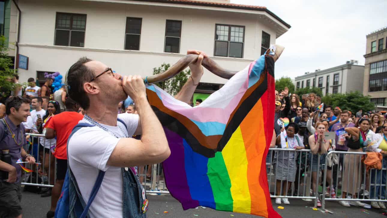man blowing horn with rainbow flag draped over it.