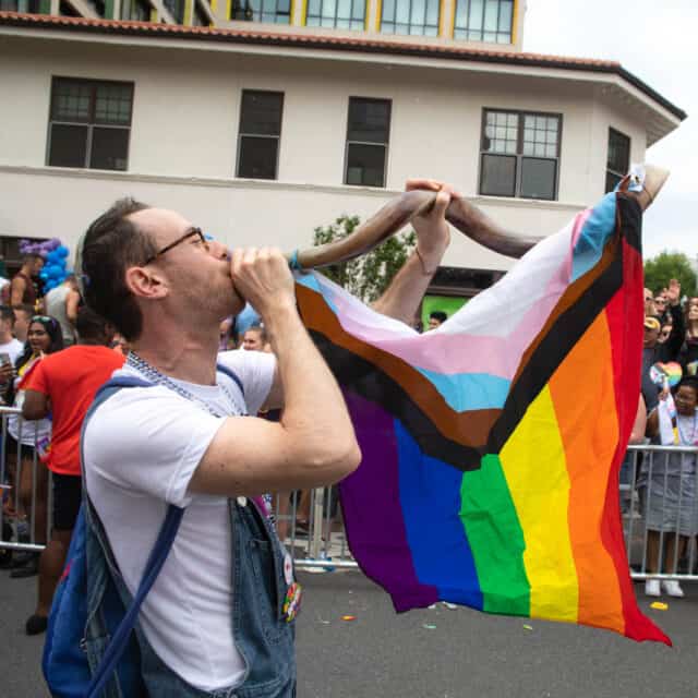 man blowing horn with rainbow flag draped over it.