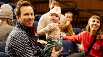 Photo of a baby in a white hat being held by their father. There are other people smiling in the background.