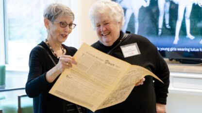Color photo of two older women, both dressed in black and with short grey or white hair, holding what looks like an old newspaper.